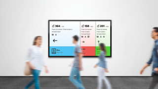 Current Lock Wireless Screendesign im Hintergrund, während Personen im Vordergrund daran vorbeilaufen.