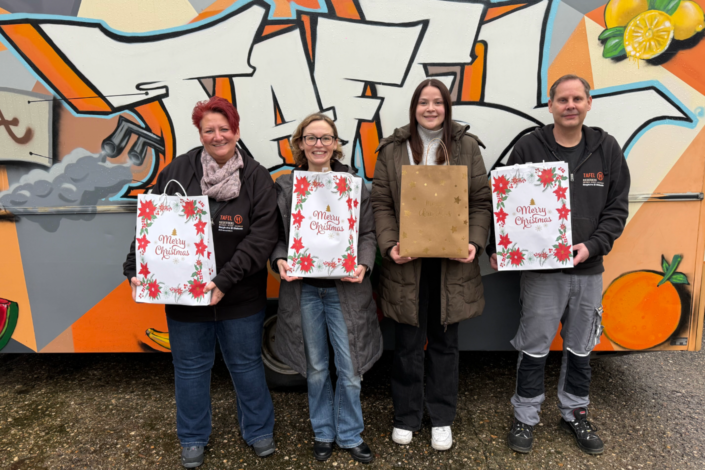 Gruppe von vier Personen mit jeweils einer Weihnachtstüte in der Hand, posieren lächelnd vor einer Graffiti-Wand.